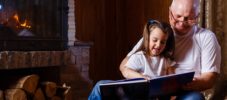 Portrait of a grandfather wearing white t-shirt reading a story to his small pretty granddaughter hugging him and listening attentively at home near the fireplace