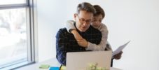 Businessman using laptop at desk in office with son embracing him