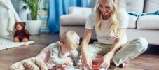 A babysitter with a 1-year-old girl is reading a children's playbook at home on the floor.