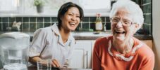 An elderly woman laughs beside a friendly young care assistant